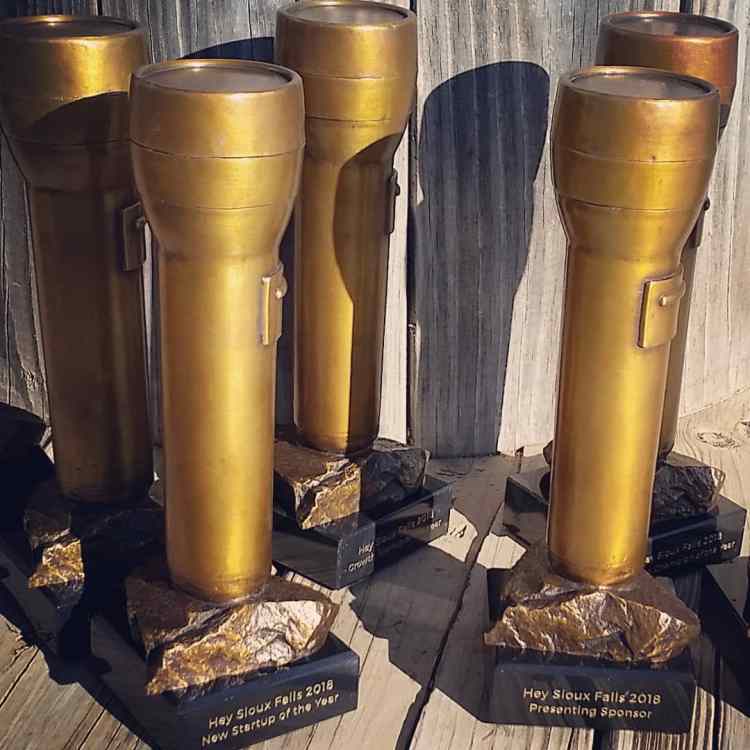 Five "Hey Sioux Falls 2018" awards. They are shaped like flashlights facing upwards with the base on a rock. They are golden in color. The front two have readable bases. The one on the left reads "Hey Sioux Falls 2018 New Startup of the Year." The one on the right reads "Hey Sioux Falls 2018 Presenting Sponsor."